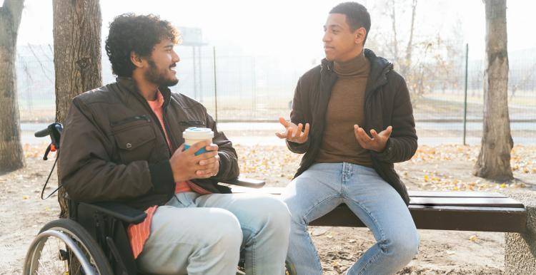 A man sits on a bench, talking to a man sitting in a wheelchair. They are outdoors and it is sunny