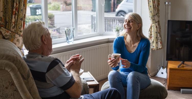 A woman sits on a footrest with a mug, talking to an elderly man in a chair